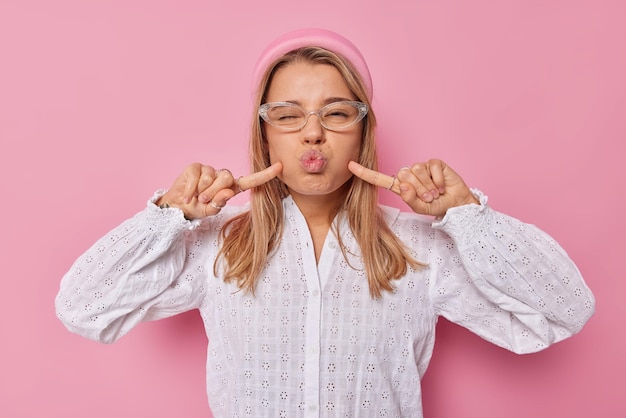 Grappig schoolmeisje dwaast rond tijdens quarantaine maakt grimas pruilend lippen en blaast wangen draagt transparante brillen stijlvolle witte blouse geïsoleerd op roze heeft speelse bui