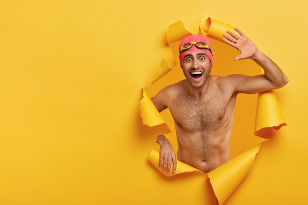 Grappige atleet man heeft een sterk naakt lichaam, golven met de handpalm, kijkt met een dolgelaatsuitdrukking