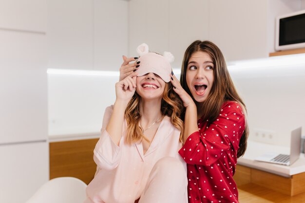 Grappige brunette meisje positieve emoties uitdrukken terwijl een grapje met vriend. Foto van prachtige blanke zusters in nachtkostuums die samen lachen.