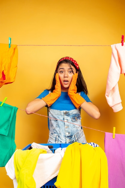 Grappige en mooie huisvrouw die huishoudelijk werk doet dat op gele ruimte wordt geïsoleerd. Jonge blanke vrouw omringd door gewassen kleren