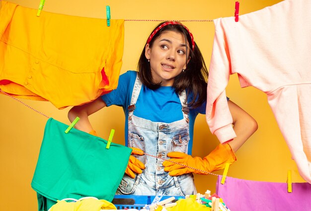 Grappige en mooie huisvrouw die huishoudelijk werk doet dat op gele ruimte wordt geïsoleerd. Jonge blanke vrouw omringd door gewassen kleren