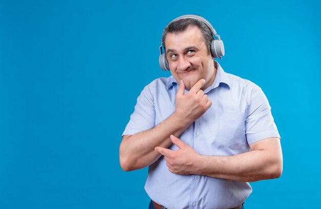 Grappige en positieve man van middelbare leeftijd in blauw gestreept shirt in koptelefoon denken en hand op kin op een blauwe ruimte