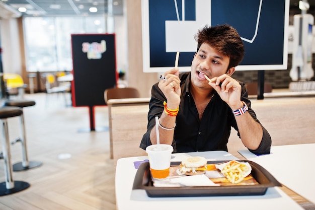 Grappige en stijlvolle Indiase man die in een fastfoodcafé zit en frietjes eet, laat zijn tong zien