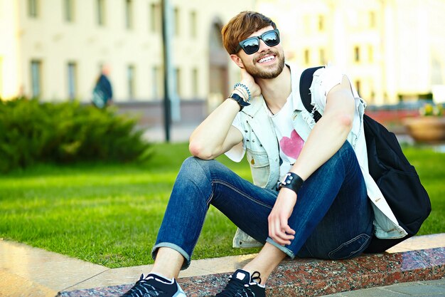 Grappige glimlachende hipster knappe mensenkerel in modieuze de zomerdoek in de straatzitting op gras in het park