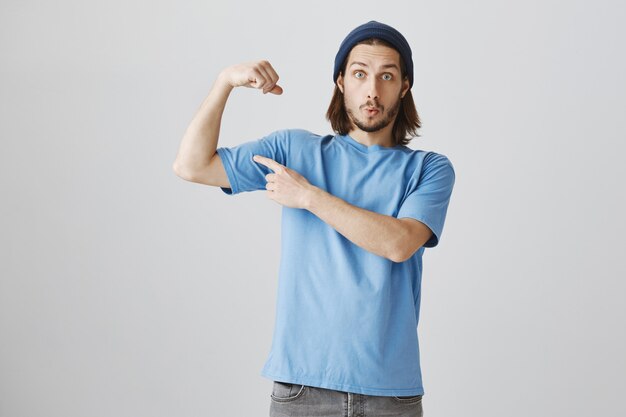 Grappige hipster man in blauw t-shirt en beanie flex biceps, sterke spieren tonen