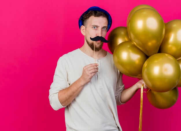 Grappige jonge knappe Slavische feestmens die feestmuts draagt ?? die ballonnen en nepsnor op stok boven de lippen houdt die naar voren kijkt en kusgebaar doet geïsoleerd op roze muur met kopie ruimte