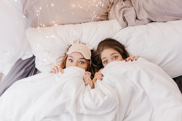 Grappige jonge meisjes verstopt onder een deken. Binnenfoto van emotionele zussen die lol hebben tijdens de ochtendfotoshoot.