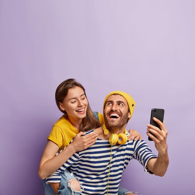 Grappige jonge paar nemen selfie op smartphone, genieten van meeliften rit, hebben gelukkige uitdrukkingen, mooie vrouw knuffels vriendje van achteren, geïsoleerd op violette achtergrond. Mensen