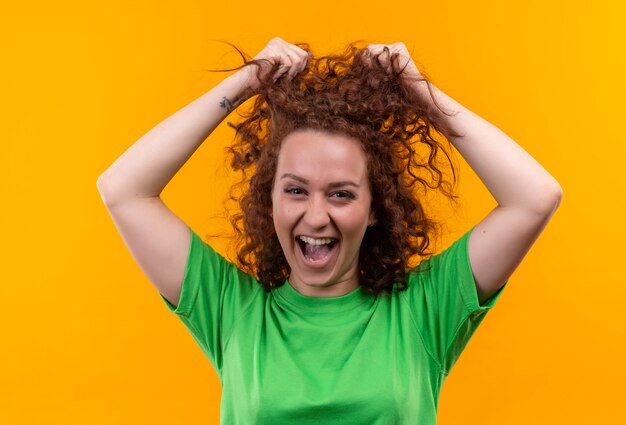 Grappige jonge vrouw met kort krullend haar in groen t-shirt camera kijken verlaten en blij haar haar aan te raken