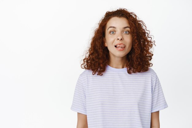 Grappige jonge vrouw met rood krullend haar, tong tonen, proberen om de neus aan te raken met het puntje van de tong, staande in t-shirt op witte achtergrond