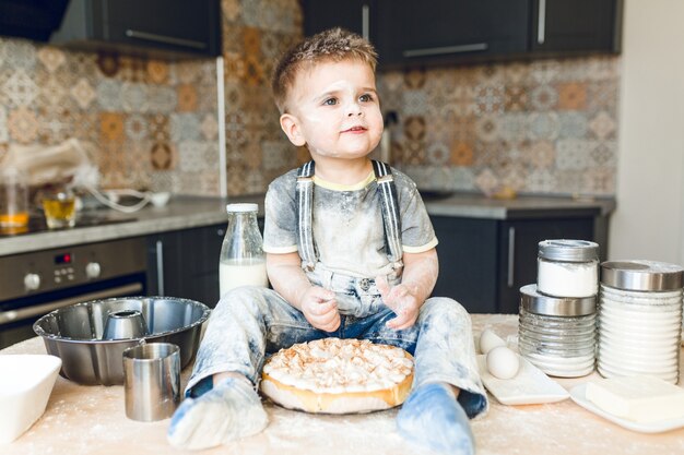 Grappige jongen zittend op de keukentafel in een akoestische keuken spelen met bloem.