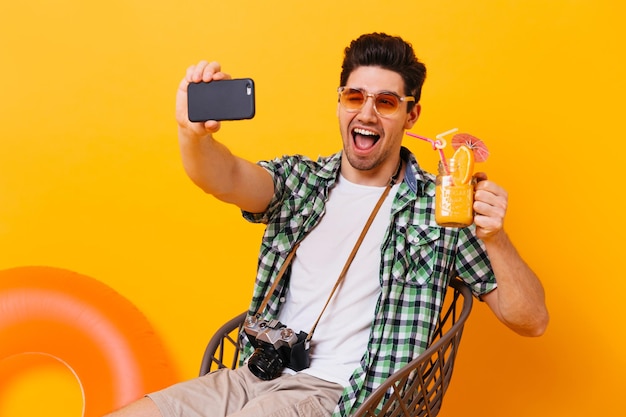 Grappige kerel in stijlvol shirt en witte t-shirt maakt selfie met een glas oranje cocktail en knipoogt op geïsoleerde achtergrond