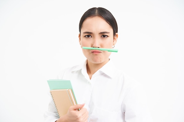 Gratis foto grappige koreaanse vrouw met notitieboekjes houdt pen met lippen en neus vast en maakt gezichten over witte stu