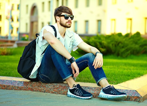 Grappige lachende hipster knappe man man in stijlvolle zomer kleding in de straat poseren zittend op gras in zonnebril