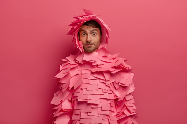 Grappige man kijkt met verwondering, trekt wenkbrauwen op, heeft geschrokken uitdrukking, draagt outfit gemaakt van zelfklevende notities, geïsoleerd over roze muur. europese man bedekt met roze stickers voor notities.