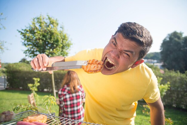 Grappige man op BBQ-familiefeest. Donkerharige man in geel T-shirt die vers geroosterd vlees eet en een grappig gezicht trekt. BBQ, koken, eten, familieconcept