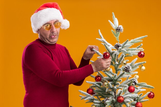 Grappige man van middelbare leeftijd met een kerstmuts in een donkerrode coltrui en een gele bril die zijn tong uitsteekt om een kerstboom te versieren die over een oranje muur staat