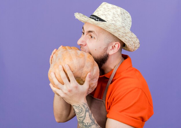 Grappige mannelijke tuinman met tuinhoed doet alsof hij pompoen bijt