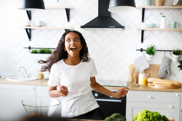 Grappige mulatvrouw die zich met volledige mond van voedsel op de moderne keuken bewegen gekleed in witte t-shirt
