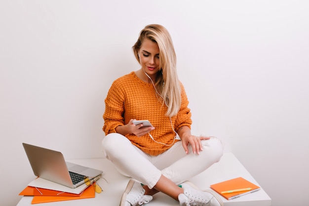 Grappige vrouw in witte sneakers zittend op het bureau met telefoon in handen en muziek luisteren