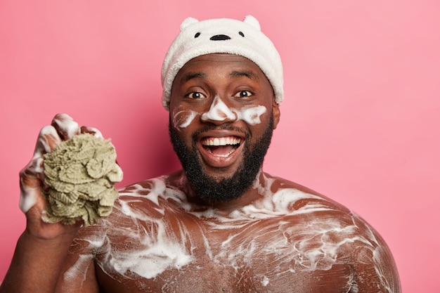Grappige zwarte bebaarde man wast zijn romp, heeft schuim op lichaam en gezicht, lacht vrolijk, houdt spons vast, draagt badmuts, geïsoleerd op roze achtergrond.