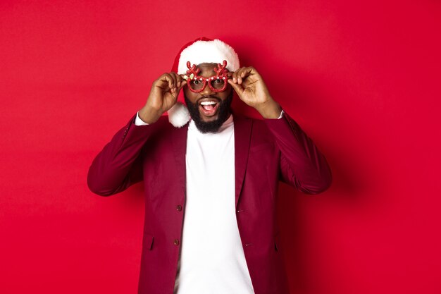 Grappige zwarte man die Nieuwjaar viert, een feestbril en een kerstmuts draagt, plezier heeft op rode achtergrond