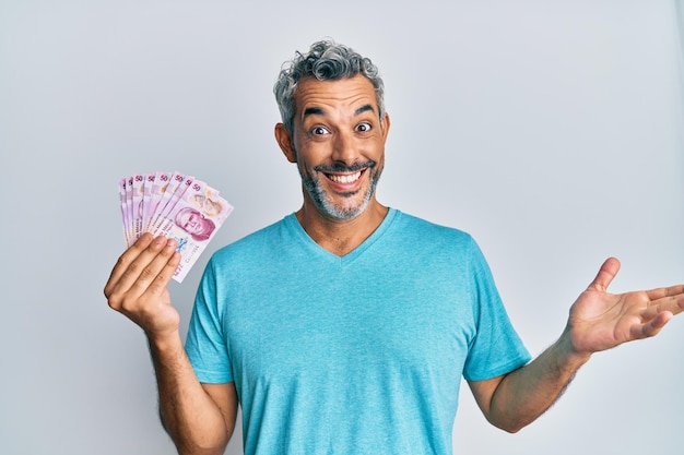 Grijsharige man van middelbare leeftijd die Mexicaanse peso's vasthoudt en prestatie viert met een gelukkige glimlach en winnaarsuitdrukking met opgeheven hand