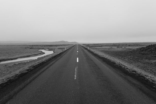 Grijstintenopname van een weg midden in een leeg veld onder een bewolkte hemel