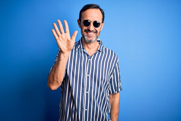 Grijze man van middelbare leeftijd met gestreept shirt en zonnebril over geïsoleerde blauwe achtergrond die toont en omhoog wijst met vingers nummer vijf terwijl hij zelfverzekerd en gelukkig glimlacht