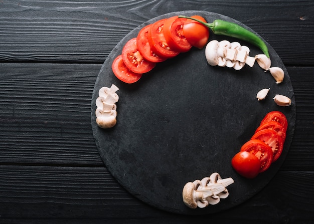 Groene chili peper; knoflookteentjes met plakjes champignons en tomaten op zwarte houten oppervlak