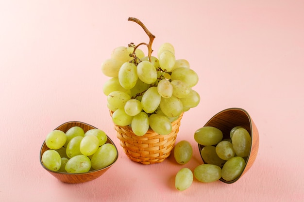 Gratis foto groene druiven in kommen en rieten mand op roze. hoge kijkhoek.