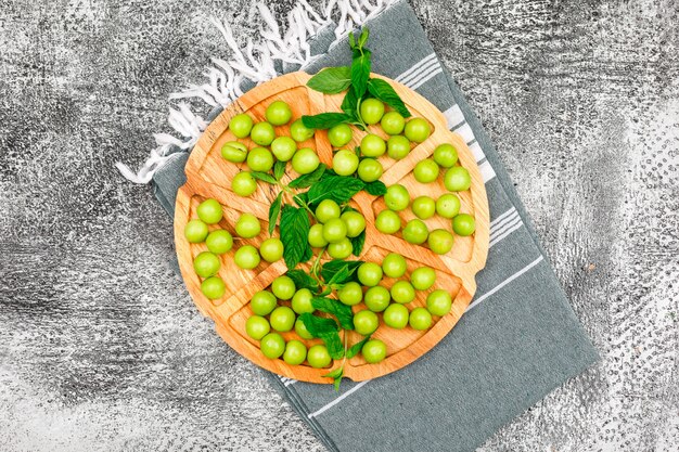 Groene pruimen met bladeren in een ronde houten plaat op grunge en picknick doek. plat lag.