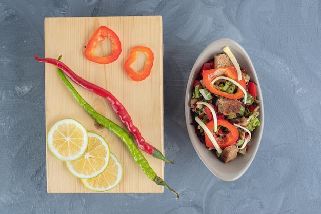 Groente salaed naast een houten bord met chili pepers en plakjes citroen en paprika op marmeren tafel.