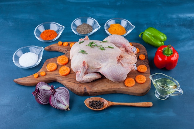 Groenten, kruiden, olie, lepel en rauwe hele kip op een snijplank op het blauwe oppervlak Gratis Foto