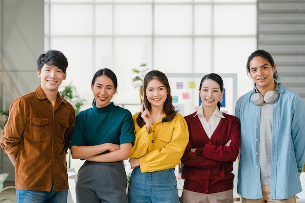 Groep Aziatische jonge creatieve mensen in slimme vrijetijdskleding glimlachen en armen gekruist in creatieve kantoorwerkplek. Diverse Aziatische mannen en vrouwen staan samen bij het opstarten. Collega teamwerk concept.