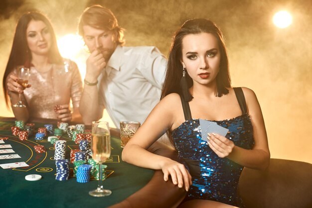 Groep elegante mensen die poker spelen in het gokhuis. Focus op een emotionele brunette in een blauwe glanzende jurk. Passie, kaarten, chips, alcohol, dobbelstenen, gokken, casino - het is entertainment. Dange