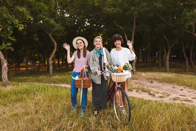 Groep gelukkige meisjes met fiets en manden vol wilde bloemen en fruit die vrolijk zwaaien en in de camera kijken in het park