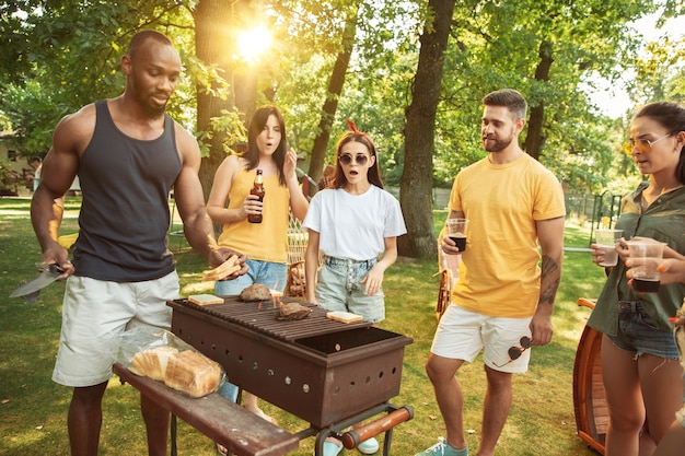 Groep gelukkige vrienden die bier en barbecuepartij hebben op zonnige dag. Samen buiten rusten in een bos open plek of achtertuin