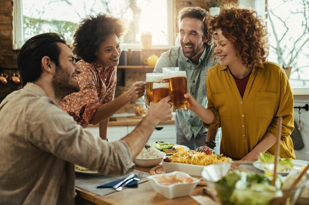 Groep jonge gelukkige mensen die plezier hebben tijdens het roosteren met bier tijdens de lunch in de eetkamer.