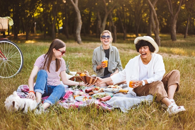 Gratis foto groep lachende meisjes die graag tijd doorbrengen op een mooie picknick met een kleine hond in het park