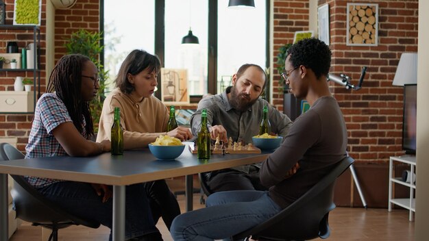 Groep mensen die nadenken over de volgende zet bij schaakbordspel, samen spelen met alcoholische dranken en snacks. Vrolijke vrienden die plezier hebben met moderne spelactiviteiten voor vrije tijd.