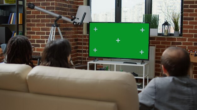 Groep mensen kijken naar groen scherm op televisie, zittend op de bank. Vrienden kijken naar chroma key-sjabloon met geïsoleerde mockup-achtergrond en lege kopieerruimte op tv. Statief geschoten.