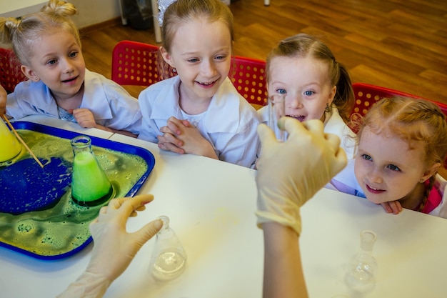 Groep schoolmeisjeskinderen met leraar in schoollaboratorium die experiment maken en de chemische reactie observeren met de kleurstof met azijn en soda-vulkaan die witte jurk uniform glas draagt