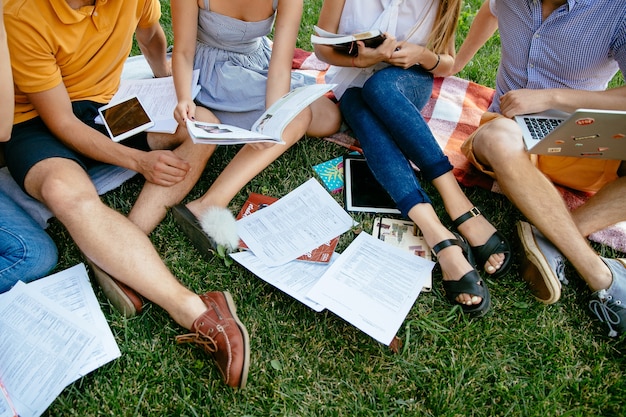 Groep studenten met boeken en tablet studeert buitenshuis samen, zittend op het gras.