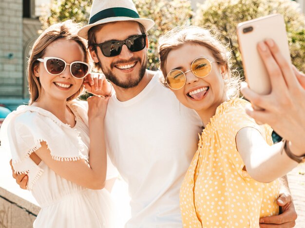 Groep van jonge drie stijlvolle vrienden in de straat. Man en twee schattige meisjes gekleed in casual zomerkleding. Lachende modellen plezier in zonnebril. Vrouwen en jongen foto selfie maken op smartphone