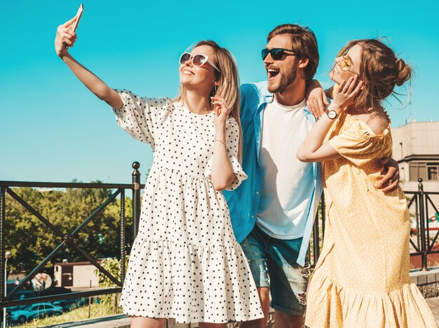 Groep van jonge drie stijlvolle vrienden in de straat. Man en twee schattige meisjes gekleed in casual zomerkleding. Lachende modellen plezier in zonnebril. Vrouwen en jongen foto selfie maken op smartphone