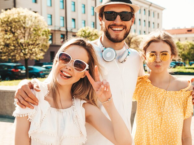 Groep van jonge drie stijlvolle vrienden poseren in de straat. Mode man en twee schattige meisjes gekleed in casual zomerkleding. Glimlachende modellen die pret in zonnebril hebben. Vrolijke vrouwen en kerel die gek worden