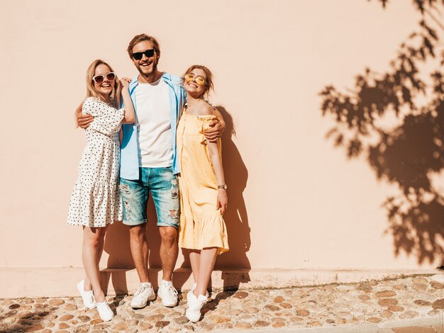 Groep van jonge drie stijlvolle vrienden poseren in de straat. Mode man en twee schattige meisjes gekleed in casual zomerkleding. Glimlachende modellen die pret in zonnebril hebben. Vrolijke vrouwen en kerel die gek worden