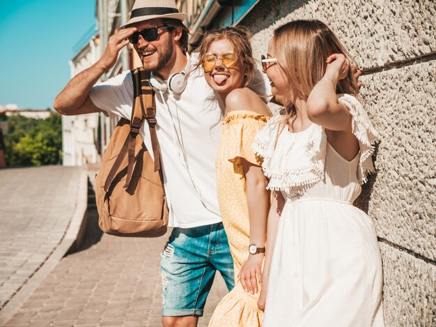 Groep van jonge drie stijlvolle vrienden poseren in de straat. Mode man en twee schattige meisjes gekleed in casual zomerkleding. Glimlachende modellen die pret in zonnebril hebben. Vrolijke vrouwen en kerel in openlucht