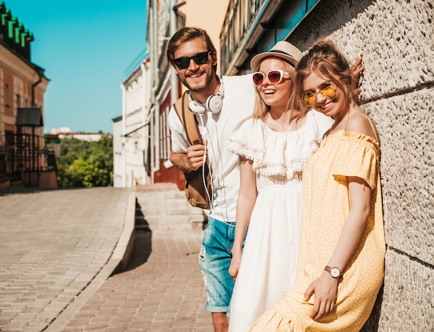 Groep van jonge drie stijlvolle vrienden poseren in de straat. Mode man en twee schattige meisjes gekleed in casual zomerkleding. Glimlachende modellen die pret in zonnebril hebben. Vrolijke vrouwen en kerel in openlucht